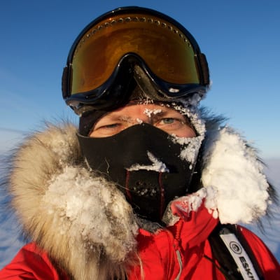 En kvinna iklädd en tjock vinterjacka med pälskantad huva, en balaklava som täcker allt utom ögonen och stora glasögon som hon för tillfället dragit upp på huvudet. Hon befinner sig på Grönland. Huvan är täckt av snö.