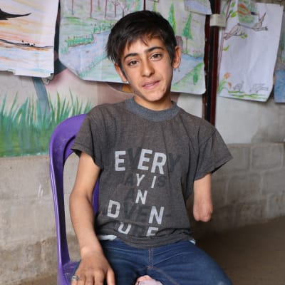 Syriska Amir sitter på en stol framför en vägg med barnteckningar. Hans armstump syns.