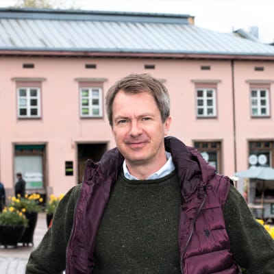 En medelålders man, tittar in i kameran med nöjd min. I bakgrunden syns ett äldre rosa stenhus och stora blomkrukor samt parkerade torgstånd och bilar. Vår eller sommar.