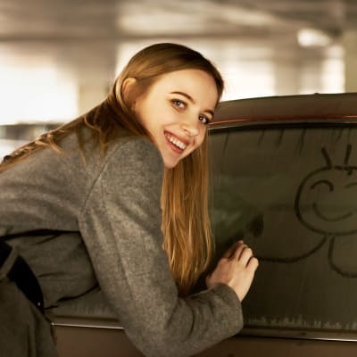 En kvinna ritar en glad gubbe på ett smutsigt bilfönster, men gubben visar mittfingret. Hon själv ler till kameran.