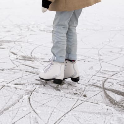 Ett barn med ljusa jeans står på isen i ett par vita skridskor.