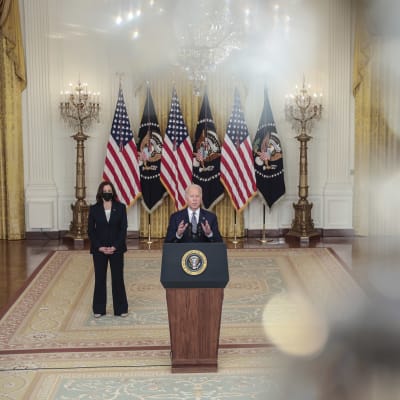 Mies seisoo puhumassa yleisölle, vierellä nainen maski kasvoillaan, taustalla Yhdysvaltojen lippuja.