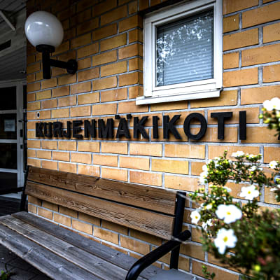 En gul tegelvägg med texten kurjenmäkikoti 2. Framför väggen finns en sliten bänk.