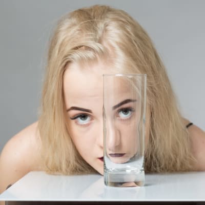Blond ung kvinna tittar mot kameran genom ettt tomt dricksglas