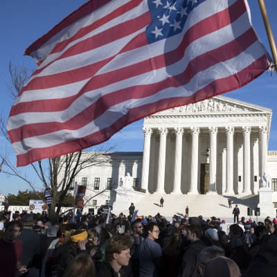 Demonstranter utanför USA:s högsta domstol en solig vinterdag. En stor amerikansk flagga vajar i förgrunden.