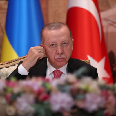 President Recep Tayyip Erdogan vid bord med blommor, med ukraisnka, turkiska och ryska flaggan i bakgrunden. 