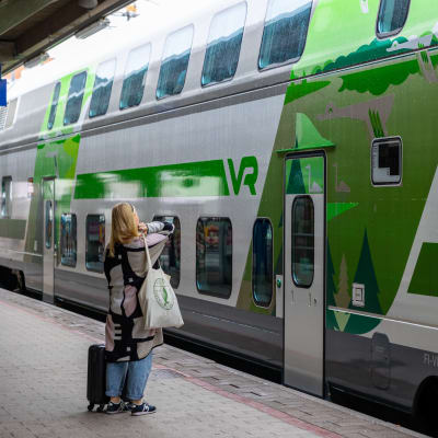 EN kvinna med resväska står på perrongen framför ett VR-tåg.