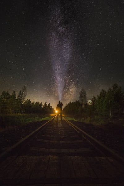 En man står på en tågbana och lyser i mörkret med en stark lampa. Stjärnor. Skog.