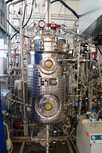 Bild inifrån ett laboratorium. I mitten står en bioreaktor, den är gjord i blänkande metall och har rör och slangar som går in i den cylindriska kroppen. 