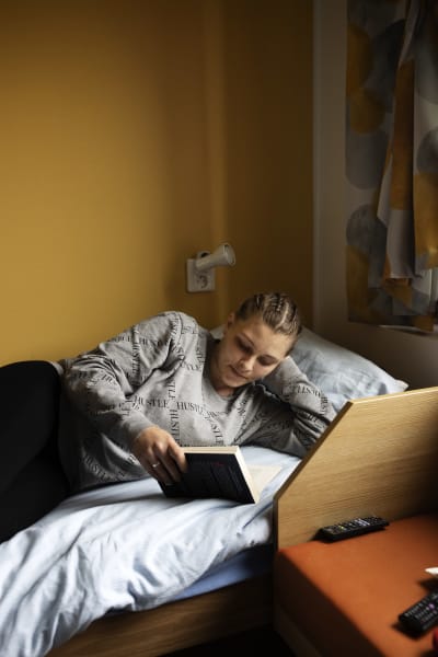 Collegepaitainen nainen makaa sängyllä ja pitää kädessään kirjaa.