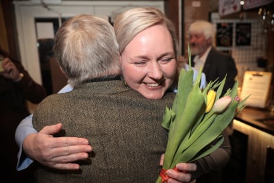 Pia Sillanpää hymyilee onnellisena tulppaanit käsissä kun häntä halaa harmaahiuksinen henkilö.