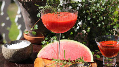 Röd drink i cocktailglas med rosmarinkvist, kumminfrön och melon i bakgrunden.