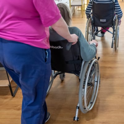 Vårdare skuffar rullstol, i vilken en äldre person sitter.