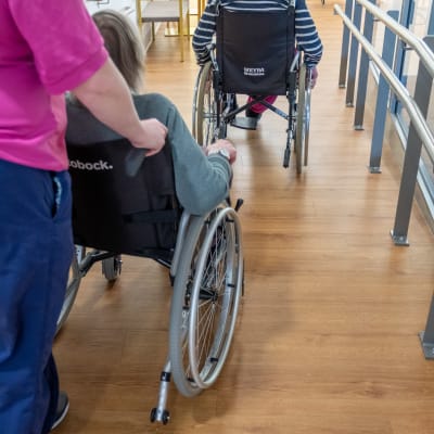 Vårdare skuffar rullstol, i vilken en äldre person sitter.