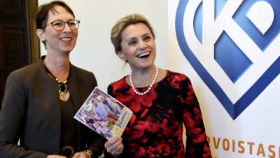 En leende Sari Essayah och en leende Päivi Räsänen, som håller i en broschyr, framför Kristdemokraternas logga.