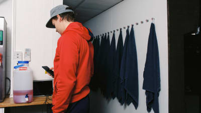 Punapaitainen jääkiekon pelaaja valitsee soitettavaa kappaletta puhelimestaan. Vieressä on mehukanisteri ja rivi pyyhkeitä seinällä.