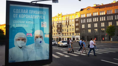 Moskvaborna uppmanas ta coronavaccin. En skylt i stadsbilden kommer med uppmaningen. I bakgrunden går folk över ett övergångsställe.