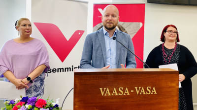 Jussi Saramo står vid ett podium i mitten, i ljusblå kavaj och skjorta, Katja Hänninen till vänster i ljuslila blus och Pia Lohikoski till höger i svart tröja och prickig klänning, bakom dem en roll-up med Vänsterns symbol "V"