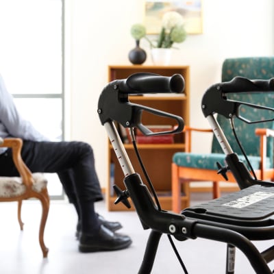 Äldre man sitter på en stol och tittar bort. I förgrunden ser man handtagen till en rollator.