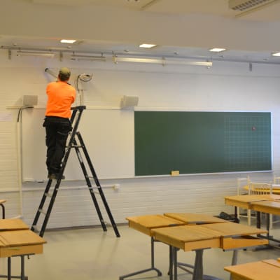 En man i orange tröja står på en stege och lägger tätning vid taklisten i ett klassrum.