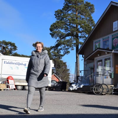 En kvinna i grå jacka går ute, i bakgrunden syns en liten bybutik och en fiskförsäljningsvagn.