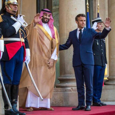 Mohammed bin Salman och Emmanuel Macron står och vinkar på Élyséepalatsets trappa. Mohammed bin Salman är klädd i rödrutig huvudduk och fotsid kaftan med gyllene dekorationer. Macron är klädd i blå kostym. Intill dem står soldater i givakt.