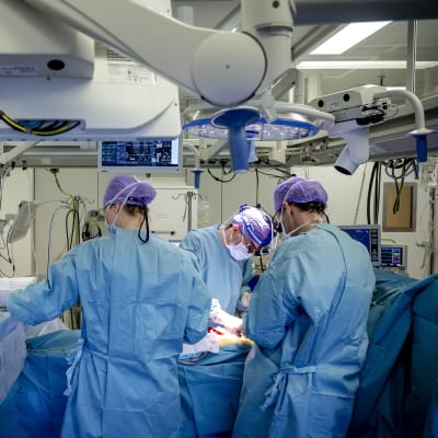 Kolme hoitajankaapuihin pukeutunutta henkilöä leikkaussalissa operoi potilasta. Huoneessa on kosolti sairaalateknologiaa.