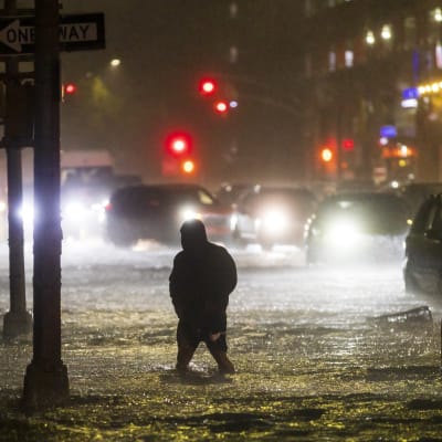 En man vadar genom en översvämmad gata i New York, upplyst av lamporna från bilar som har fastnat i vattenmassorna.