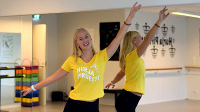 En flicka står framför spegeln i en danssal med armarna utsträckta. Hon ler och har på sig en gul t-skjorta med texten Hurja Piruetti.