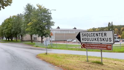 En vit vägskylt med texten Skolcenter Koulukeskus, och en brun skylt med texten Pargas hembygdsmuseum, i en vägkorsning med en skolbyggnad i bakgrunden.