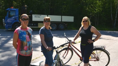 Samira Jaakkola, Sari Siikala och Kaarina Hurtta står vid Eriksnäsvägen. Siikala och Hurtta håller i cyklar. Bakom kör en stor lastbil.