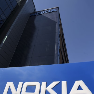 Stor skylt med namnet Nokia i Bakgrunden kontorsbyggnader och blå himmel och ett moln fotograferade nedifrån. Nokias huvudkontor i Esbo.