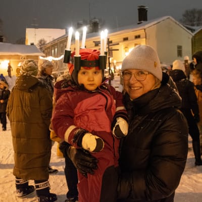 En liten flicka med luciakrona i famnen på en kvinna. Båda ler och tittar in i kameran. 