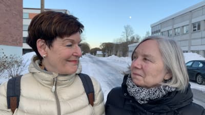 Två kvinnor står tillsammans utomhus på en trottoar, det är vinter och snö på marken. Kvinnorna tittar på varandra och ler.