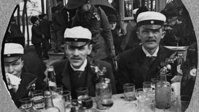 En svartvit gammal bild på tre män som i studentmössor som sitter vid ett bord utomhus med många urdruckna glas framför sig.
