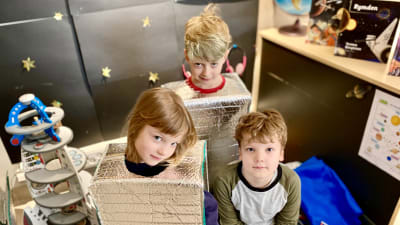 Förskoleeleverna Clara Stegar, Wilmer Åsvik och Erik Lingonblad sitter i en rymdhörna på förskolan. Clara och Wilmer är klädda i hemgjorda rymddräkter. 