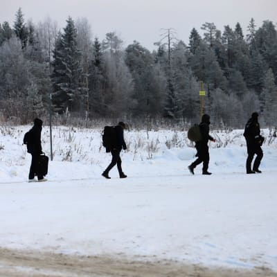 Kolme tummiin pukeutunutta ihmistä kantaa laukkuja jonossa talvisessa maisemassa, jonon kummassakin päässä kävelee rajavartija.