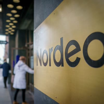 Nordean logo rakennuksen ulkoseinustalla. valkoiiseen takkiin pukeutunut epäterävä naishahmo avaa ovea, taustalla muita ihmisiä epäterävinä.