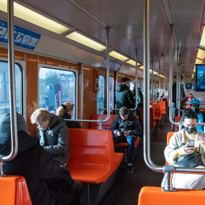 Passagerare sitter och står i en metrovagn med orangefärgade bänkar. Många eller alla ser ut att ha munskydd.