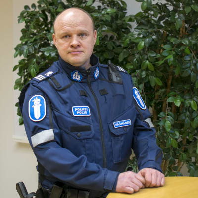 En polis står vid ett bord, han ser in i kameran och ser allvarlig ut. 