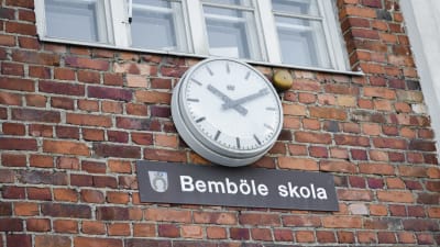 Klocka på vägg, under den skylt med texten Bemböle skola.