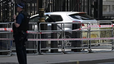 En silverfärgad personbil står intill en svart grind, omgärdad av kravallstaket och polisens plastband. En polis står en bit ifrån bilen.