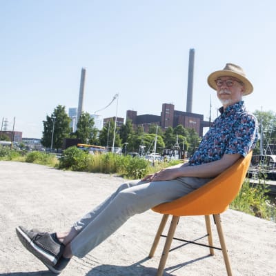Ydinvoiman mallinnuksen professori Juhani Hyvärinen istuu oranssilla tuolilla Hanasaaren voimalan edustalla.