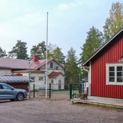 En gård med två trähus, ett rött och ett gult. En bil står parkerad vid en flaggstång.