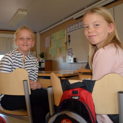 Två flickor sitter i ett klassrum vid sina pulpeter.