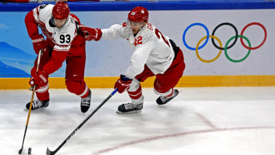 En dansk hockeyspelare med röd tröja har pucken mot en rysk spelare i vit tröja utan puck.