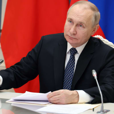 Vladimir Putin sitter vid ett bord framför den ryska flaggan. På bordet finns stående mikrofoner och papper.