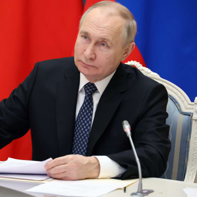 Vladimir Putin sitter vid ett bord framför den ryska flaggan. På bordet finns stående mikrofoner och papper.
