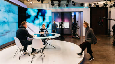 Two persons sitting at a white table in a tv studio in front of a monitor wall. 