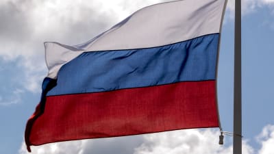 Den ryska flaggan vajar i vinden med blå himmel och några moln i bakgrunden.